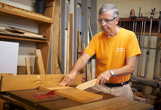 Older white male cutting a piece of wood using a table saw.